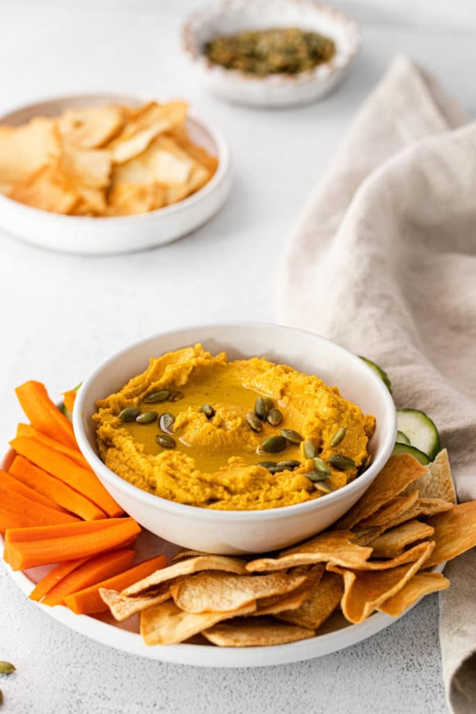 A bowl of pumpkin hummus served with pita chips, carrot sticks and cucumber slices.
