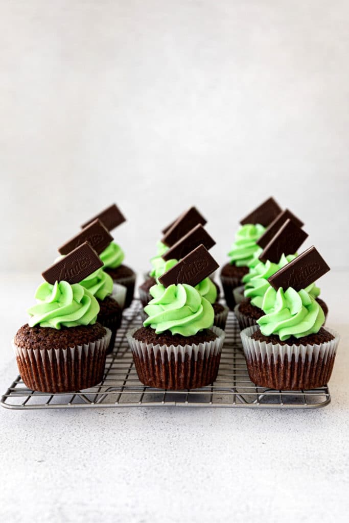 One dozen mint chocolate cupcakes with mint frosting, garnished with Andes mints, on a wire rack.