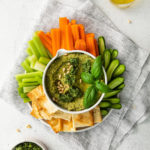 Overhead view of basil pesto hummus in a small bowl garnished with pine nuts, surrounded with veggies and crackers.