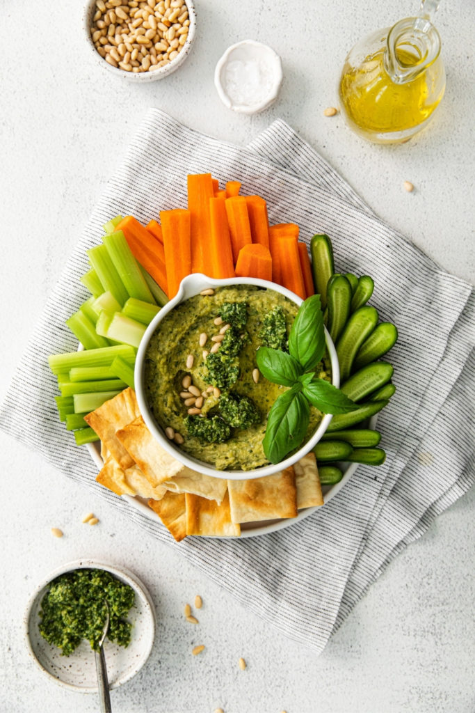 Overhead view of basil pesto hummus in a small bowl garnished with pine nuts, surrounded with veggies and crackers.