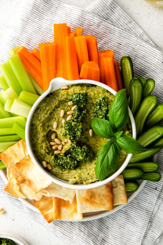 Pesto hummus appetizer on a table with buttery crackers, carrot and celery sticks, and baby cucumbers.