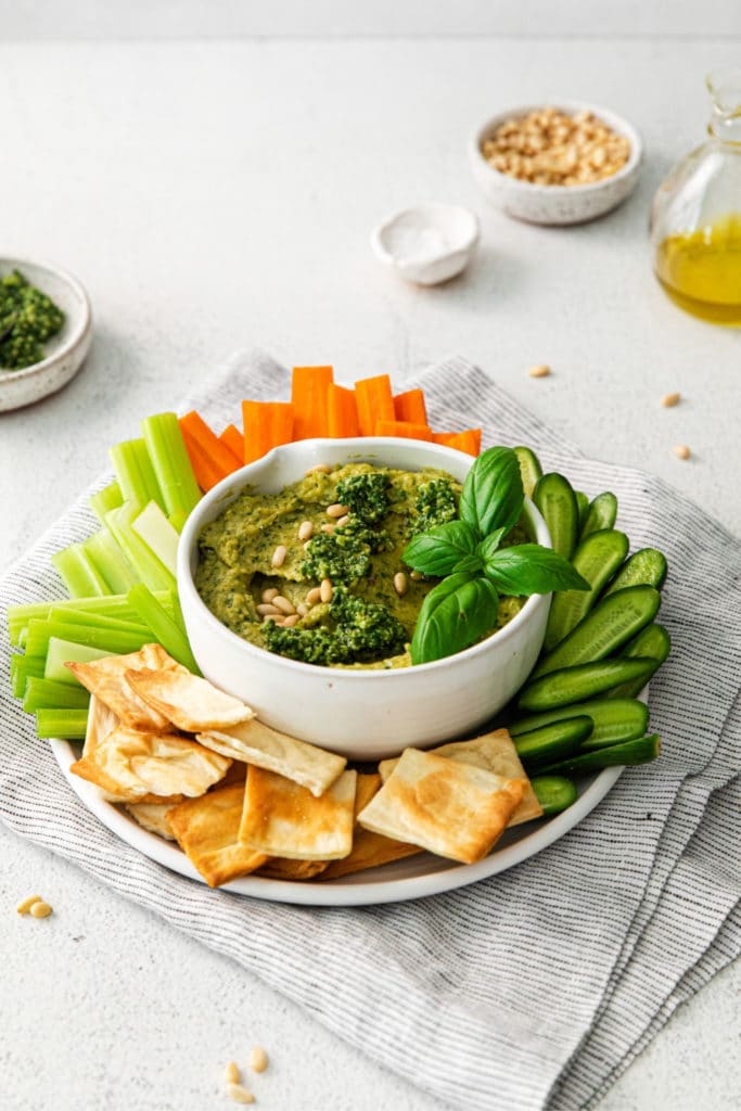 Pesto Hummus with basil served in a small appetizer bowl with crackers and veggies.