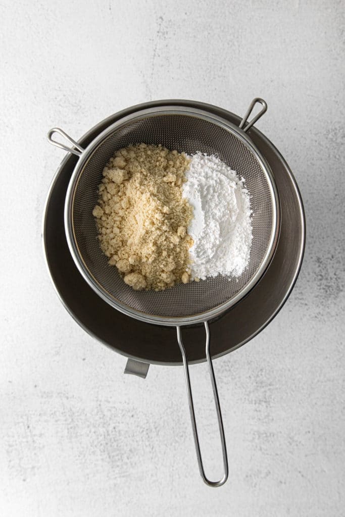 Almond flour and powdered sugar being sifted through a fine-mesh strainer into a metal bowl.