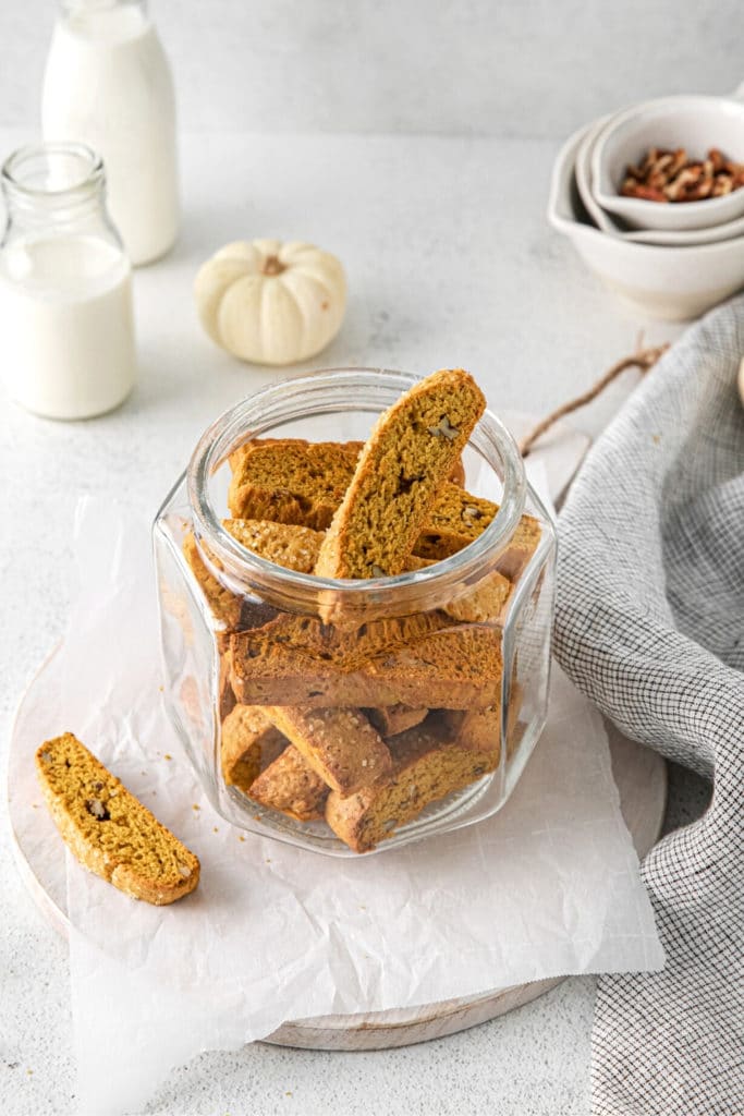 Glass cookie jar filled with homemade pumpkin biscotti Italian cookies on a dessert table with milk.