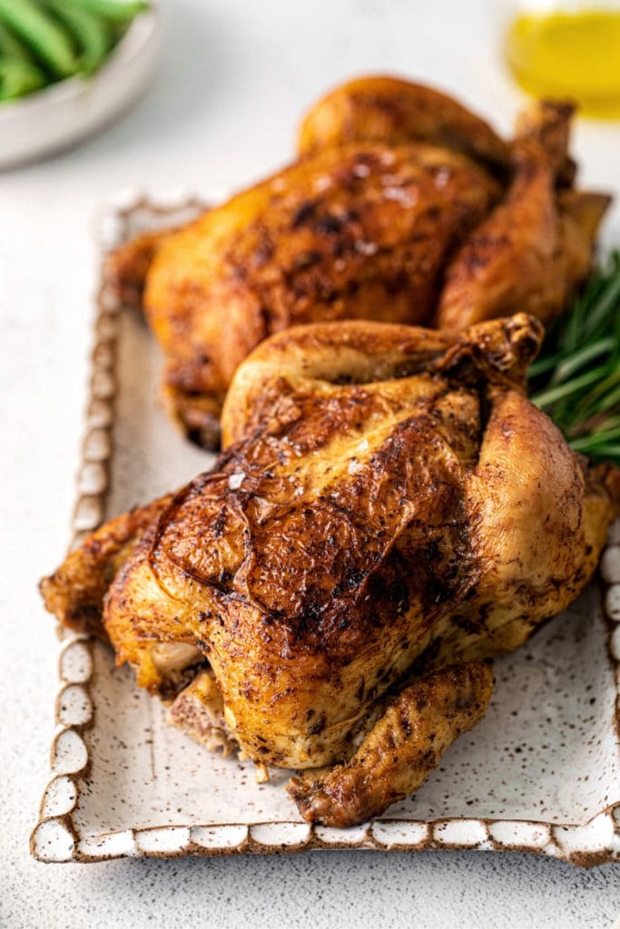 Two baked Cornish hens with crispy golden skin on a serving platter with rosemary sprigs.