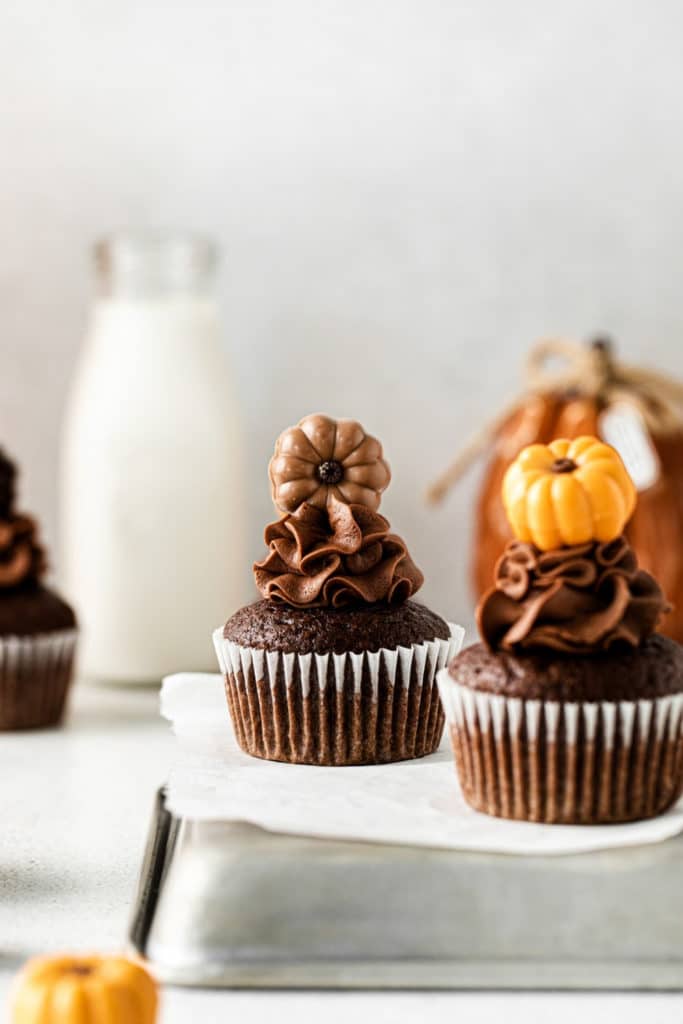 Two pumpkin chocolate cupcakes on a metal tray with a bottle of milk in the background.