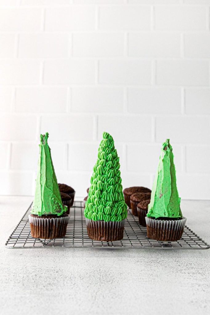 Three partially decorated Christmas tree cupcakes on a metal rack.