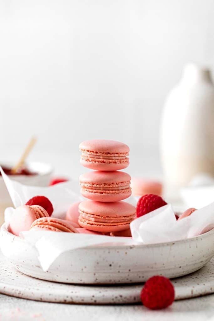 Pink valentines macarons with raspberry filling presented in a wide bowl with fresh raspberries.