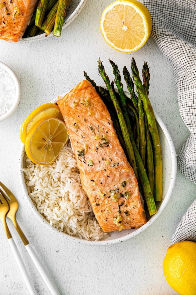 Individual serving of baked salmon with lemon butter and asparagus in a bowl with sliced lemon.