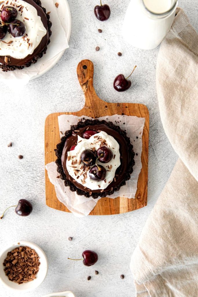 Overhead view of a chocolate tart garnished with whipped cream, chocolate shavings and dark-red cherries.