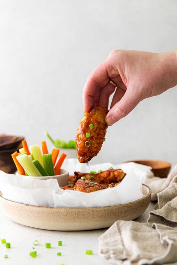 Fingers gripping and lifting a BBQ chicken wing from a serving bowl on an appetizer table.