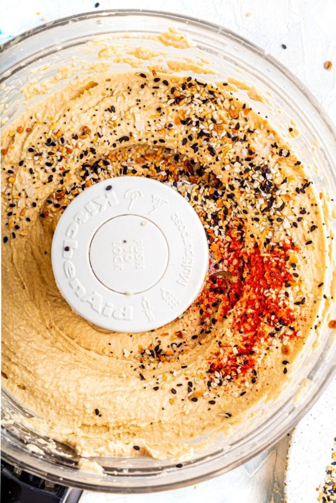 Homemade hummus in a food processor sprinkled with everything bagel seasoning and cayenne pepper.