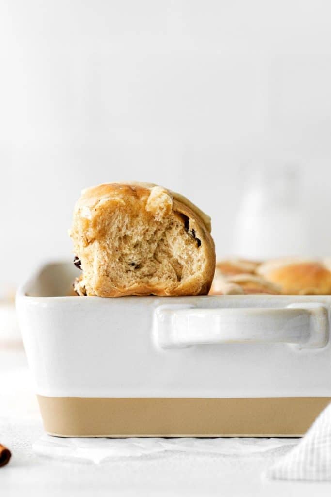 Side view of a hot cross bun set on the edge of a white ceramic baker.