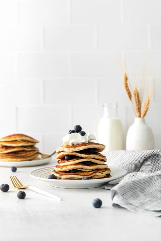 Stack of lemon blueberry pancakes on a plate next to a gold fork and glass bottle of milk.
