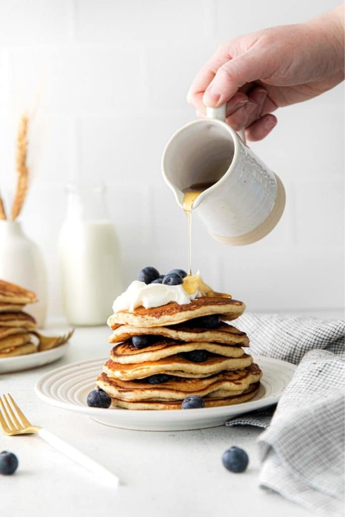 Maple syrup being poured over a stack of lemon blueberry pancakes.