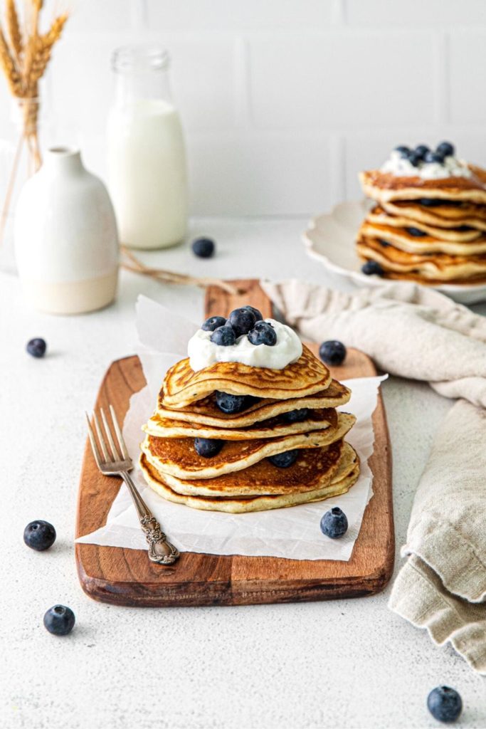Six fluffy blueberry pancakes stacked on a wooden board along with a metal fork.