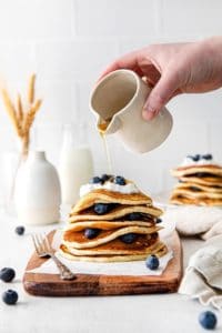 Maple syrup being drizzled over a warm stack of lemon blueberry hotcakes.