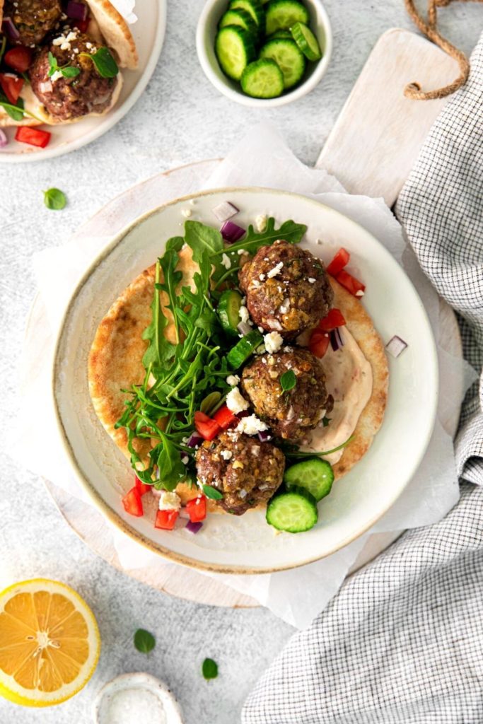Assembling a homemade meatball gyro sandwich with traditional spreads and fresh produce.