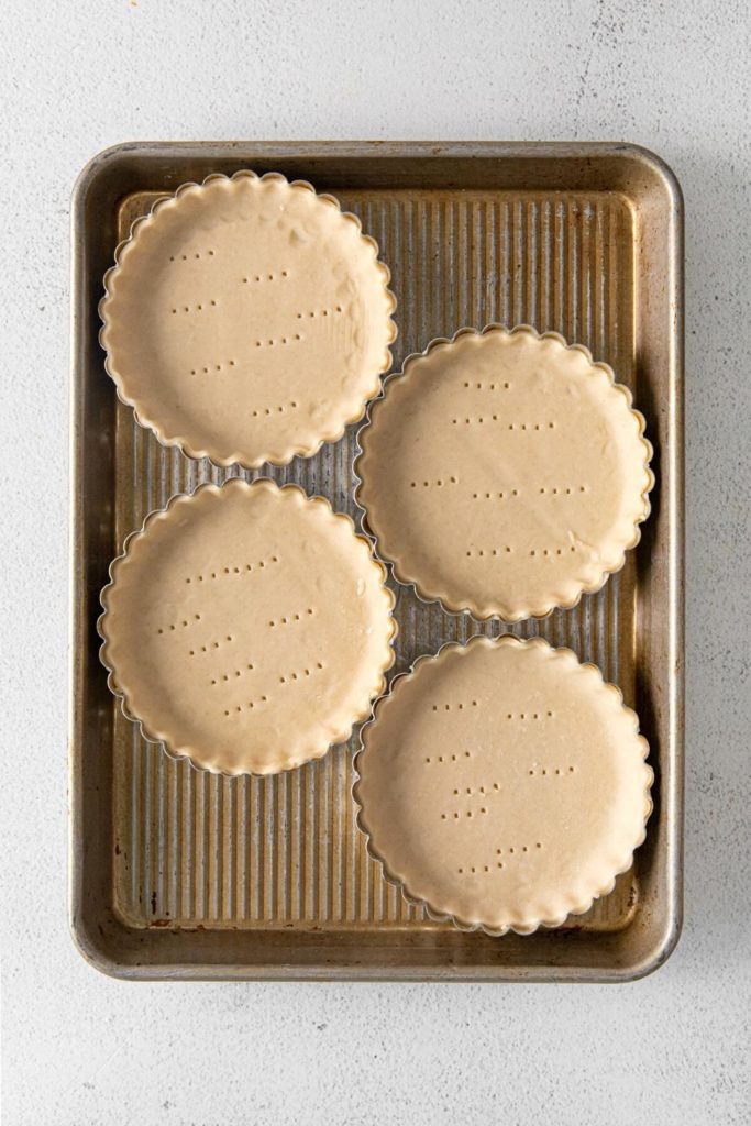 Four mini tartlet pans with raw tart dough ready to bake in the oven.