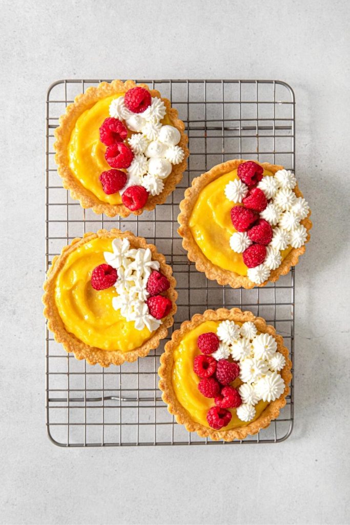 Four mini lemon tarts garnished with whipped cream and raspberries set on a metal rack.
