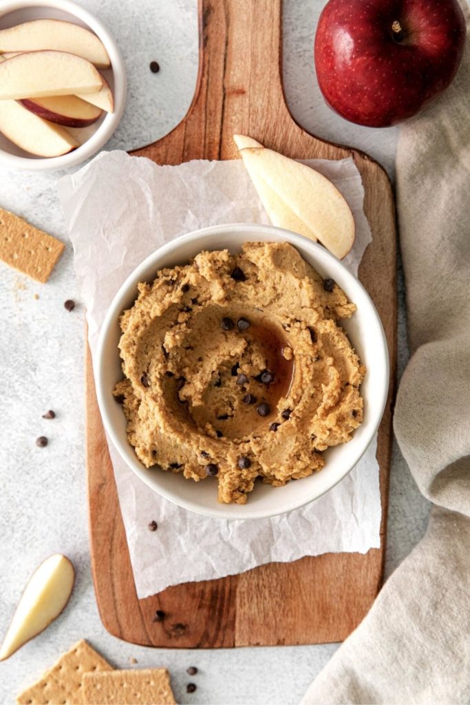 Chickpea cookie dough hummus with maple syrup swirled over the surface served with apple slices.