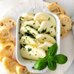 Marinated cheese slices in an appetizer dish with pieces of sliced baguette.