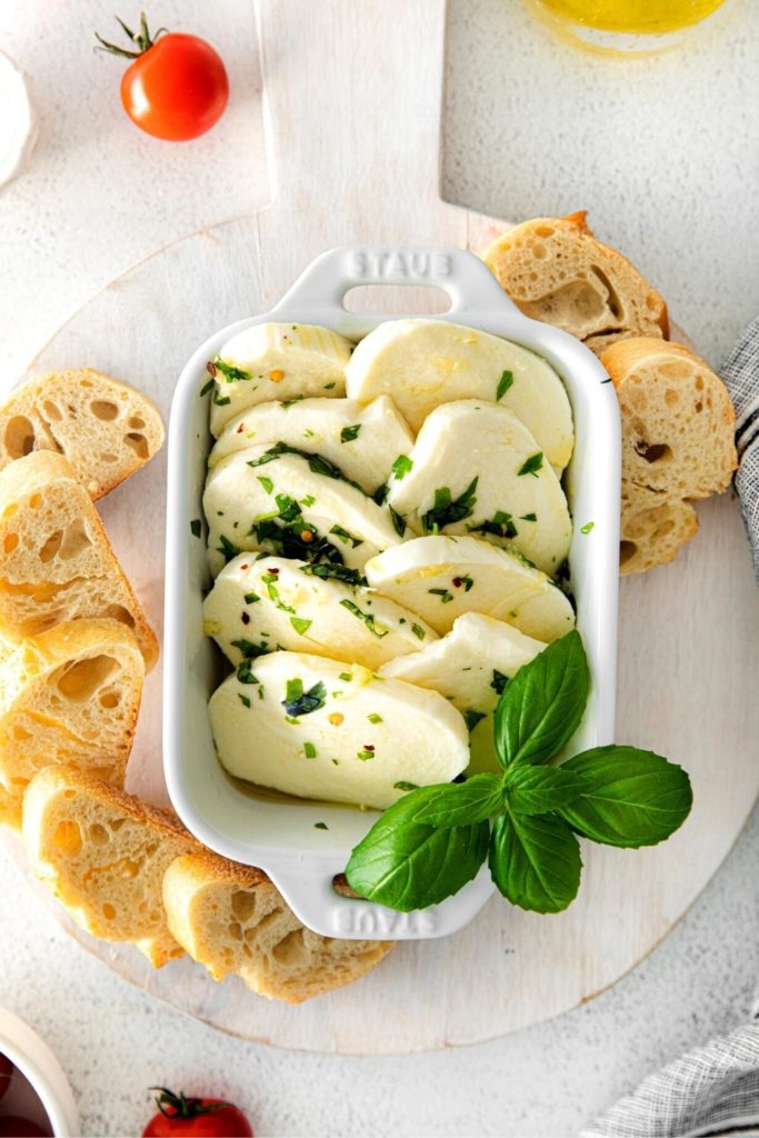 Marinated cheese slices in an appetizer dish with pieces of sliced baguette.