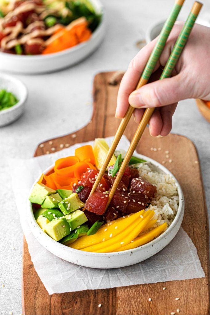 Chopsticks held in a hand lifting up a piece of ahi tuna from a spicy tuna poke bowl.