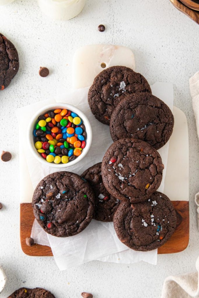 Freshly-baked chocolate M&M cookies cooling on a board alongside a bowl of M&Ms.