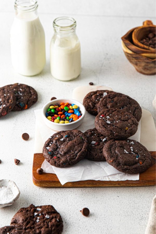 A fresh batch of chocolate M&M cookies on a serving board with a bowl of M&Ms and glass bottles of milk.