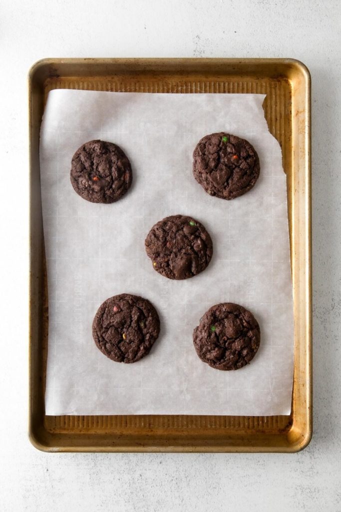 Five chocolate cookies with M&M's on a baking tray lined with parchment paper.