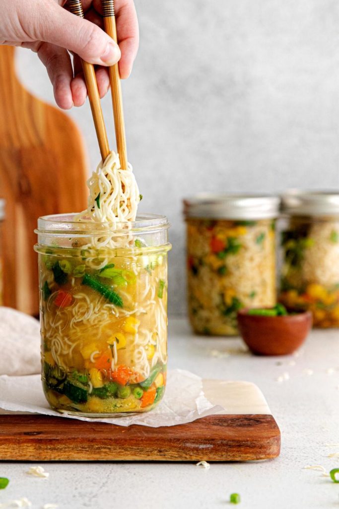 Hand with chopsticks lifting ramen noodles from a mason jar of homemade cup noodles with veggies.