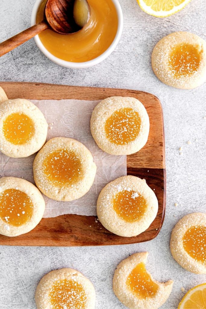 Lemon thumbprint cookies cooling on a wooden board next to a bowl of lemon curd.