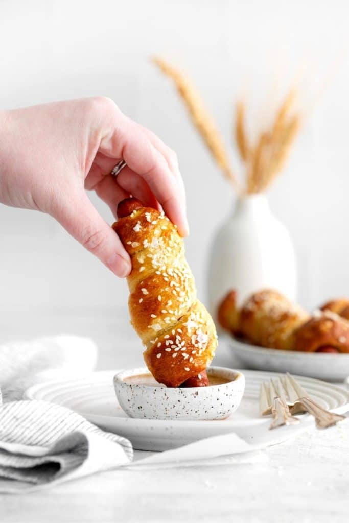 A hand holding a homemade Nathan's pretzel dog while dipping it into a dish of mustard.