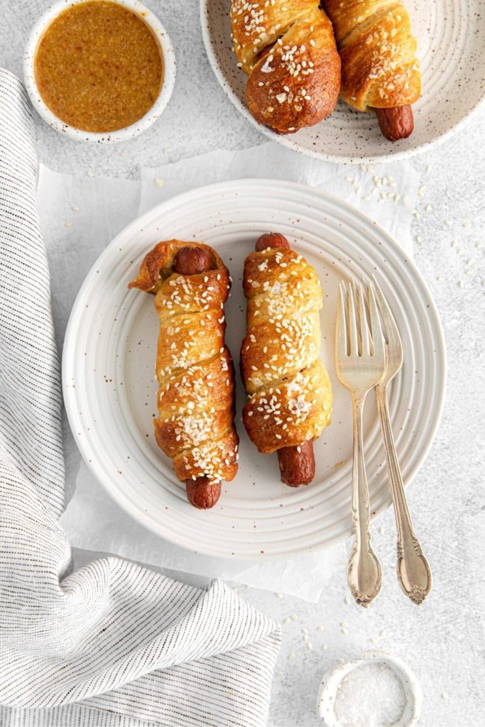 Two homemade pretzel dogs on a white lunch plate with silver forks.