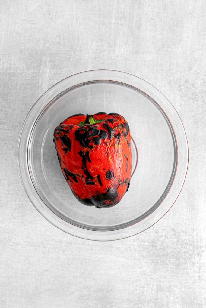 A whole fire-roasted red bell pepper in a glass bowl.