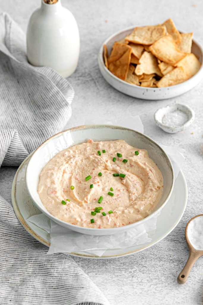 Red pepper feta dip in an appetizer bowl next to pita chips and a pinch bowl of salt.