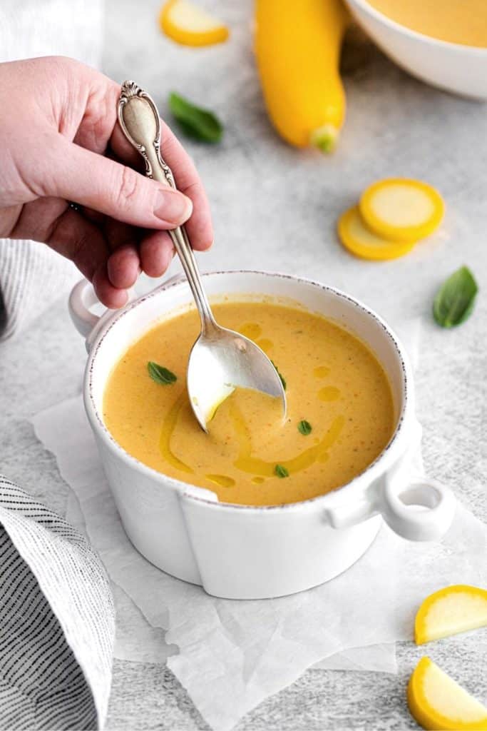 A hand holding a spoonful of yellow summer squash soup over a white soup bowl.