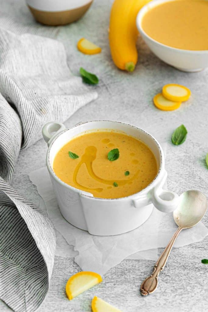 Yellow squash soup served in a white-handled soup bowl garnished with a swirl of olive oil and fresh oregano leaves.