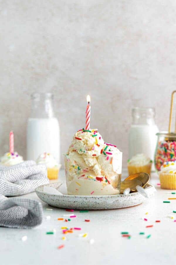 Two scoops of birthday cake ice cream in a small glass dessert cup with lit birthday candle.