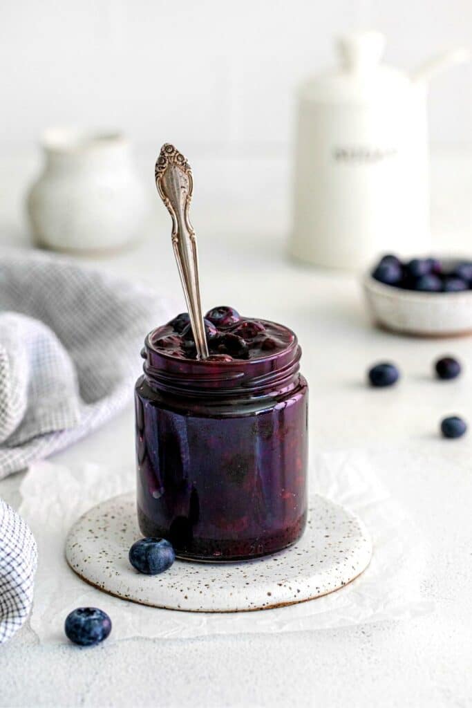 Blueberry filling in a glass jar with spoon on a serving table with fresh blueberries.