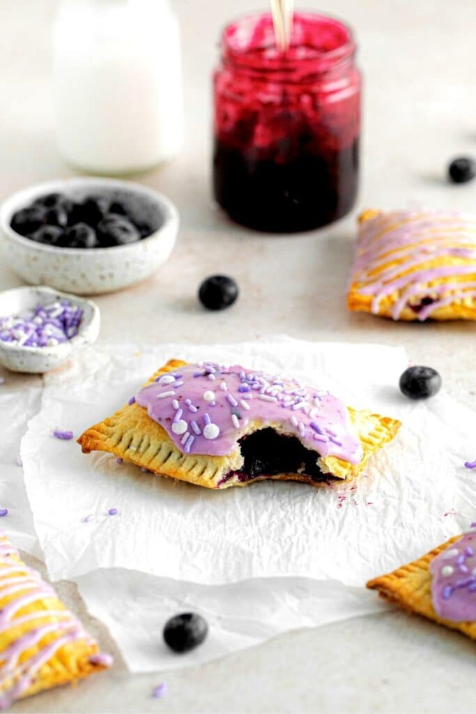 A frosted blueberry hand pie with a bite taken out on a dessert table with purple sprinkles and blueberries.