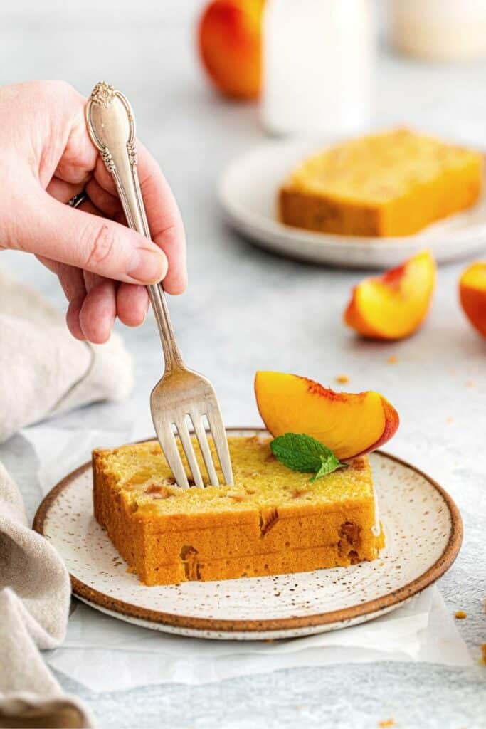 A hand with fork digging into a thick slice of peach cobbler pound cake served on a stoneware plate.