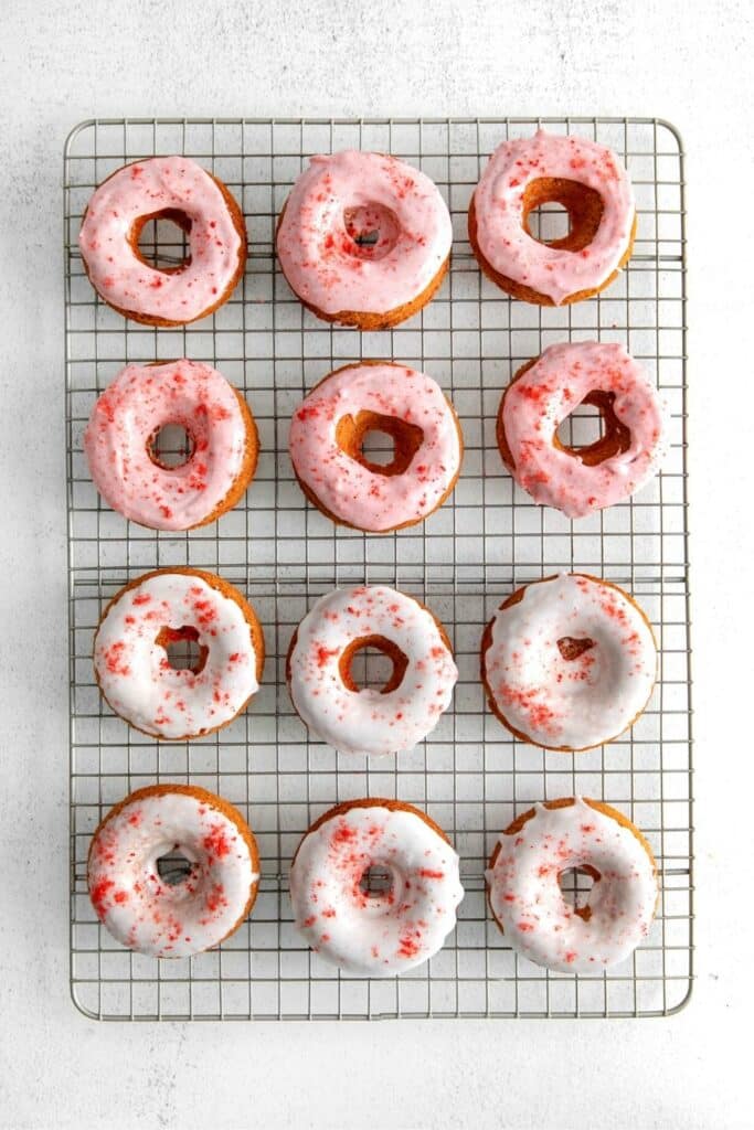 One Dozen baked strawberry donuts with pink and white glaze on a wire rack.