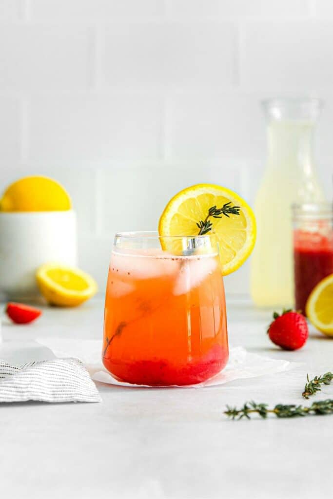 Glass of homemade pink lemonade with fresh strawberries and lemons on a white countertop.