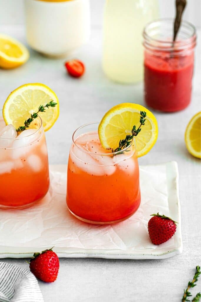 Two glasses of blended strawberry lemonade on a serving tray with a jar of strawberry syrup and a carafe of lemonade in the background.