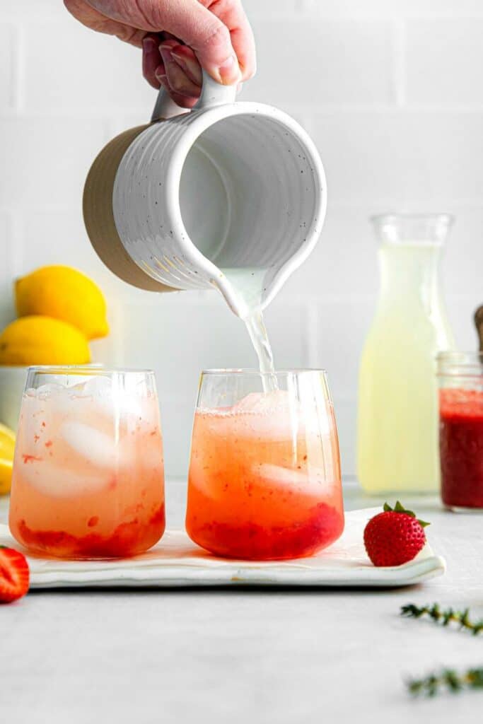 Lemonade being poured from a pitcher into a glass filled with strawberry syrup and ice.