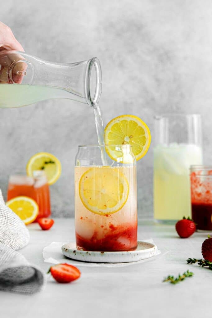 Hand pouring lemonade into an iced down glass with strawberry syrup and 2 slices of lemon.