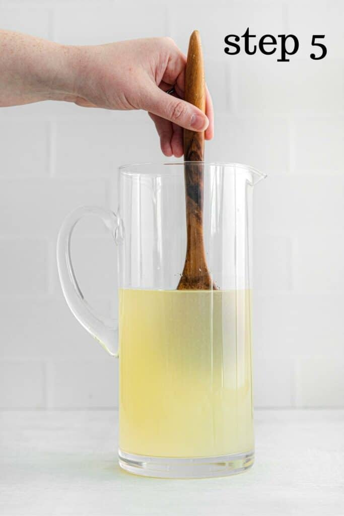 Hand stirring a tall glass pitcher of lemonade with a wooden spoon.