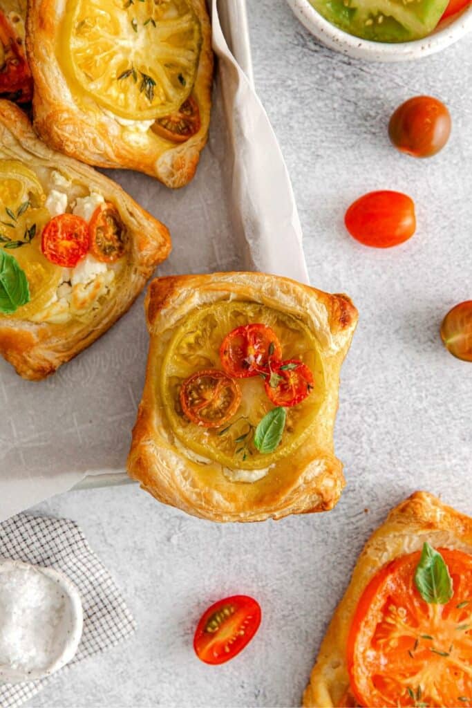 Puff pastry tomato tartlets on a serving table with fresh herbs and cherry tomatoes.